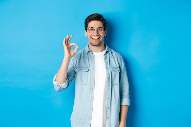 Atractivo joven con gafas y ropa casual, mostrando una buena señal de aprobación, como algo, de pie contra el fondo azul.