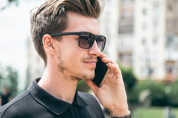 Atractivo joven con gafas de sol hablando por un teléfono inteligente en la ciudad