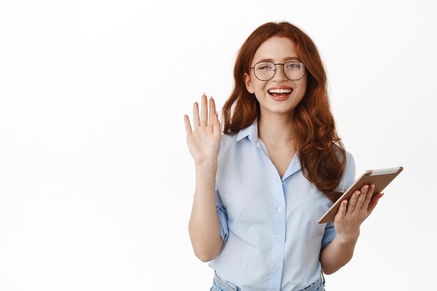 Atractivo joven gerente de oficina, compañera de trabajo de la directora ejecutiva saludando con la mano, sosteniendo una tableta digital y sonriendo amigablemente a la cámara, saludando a los clientes, de pie contra un fondo blanco.