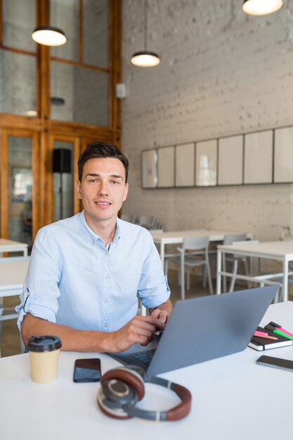 Atractivo joven guapo sonriente sentado en la oficina de espacios abiertos trabajando en equipo portátil