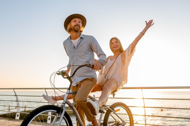 Atractivo joven hombre y mujer viajando en bicicleta, pareja romántica en vacaciones de verano junto al mar al atardecer, traje de estilo boho hipster, amigos divirtiéndose juntos