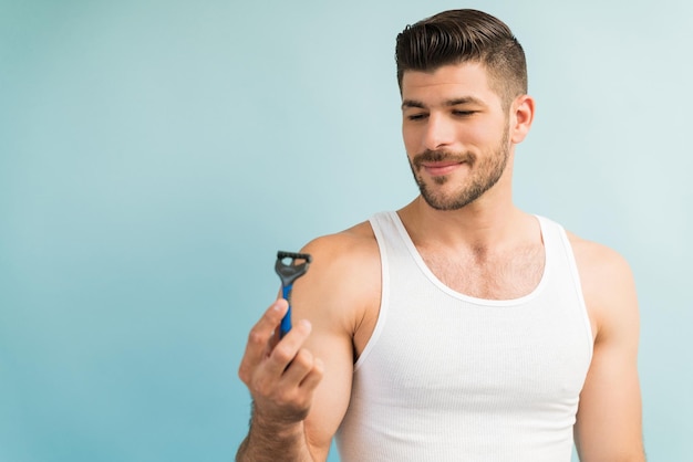 Atractivo joven latino mirando la maquinilla de afeitar con una sonrisa en el estudio