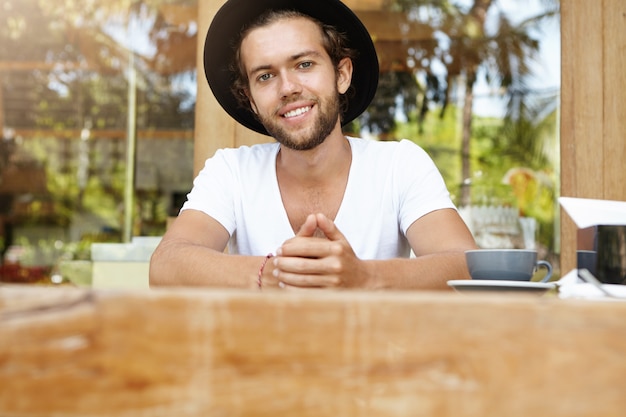 Atractivo joven de moda en sombreros de moda sentado en la mesa de madera de la cafetería.