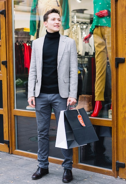 Un atractivo joven de pie fuera de la tienda con bolsas de compras en la mano