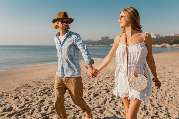 Atractivo joven sonriente feliz con sombrero y mujer rubia en vestido blanco corriendo juntos en la playa