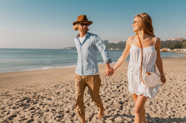 Atractivo joven sonriente feliz con sombrero y mujer rubia en vestido blanco corriendo juntos en la playa