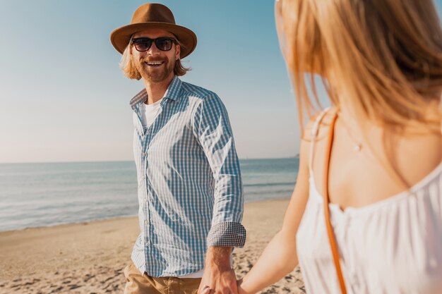 Atractivo joven sonriente feliz con sombrero y mujer rubia en vestido blanco corriendo juntos en la playa