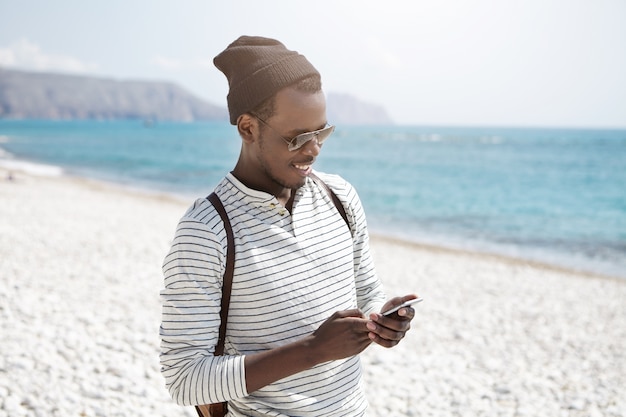 Atractivo joven viajero masculino negro sonriente en tonos de moda usando un teléfono inteligente, enviando un correo electrónico a sus familiares, luciendo feliz mientras camina por el océano solo. Personas, estilo de vida y viajes.