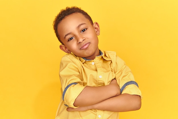 Atractivo lindo niño afroamericano fresco vestido con camisa amarilla cruzando los brazos sobre el pecho y mirando a la cámara con una sonrisa alegre, postura que expresa confianza