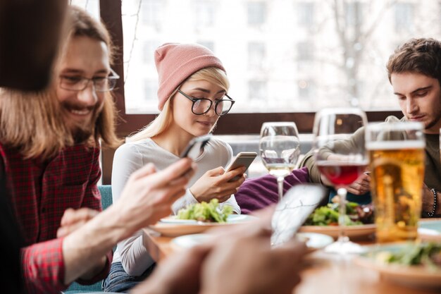 Atractivos amigos sentados en la cafetería y utilizando teléfonos móviles.