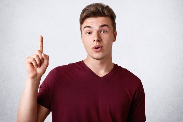 Aturdido adolescente masculino con expresión sorprendida mantiene el dedo índice levantado