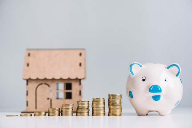 Aumentando las monedas apiladas; modelo de piggybank y de la casa en el escritorio reflexivo blanco