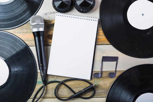 Auricular; micrófono; Disco de vinilo y cuaderno de espiral en blanco con nota musical en mesa de madera.