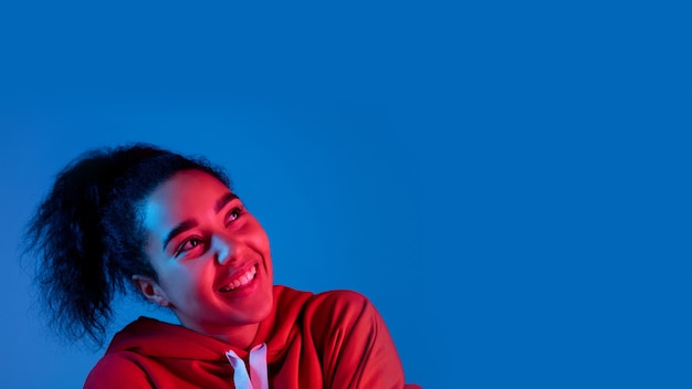 Auriculares. Retrato de mujer afroamericana aislado sobre fondo azul de estudio en luz de neón multicolor. Modelo de mujer hermosa. Concepto de emociones humanas, expresión facial, ventas, publicidad, moda.