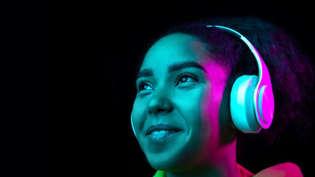 Auriculares. Retrato de mujer afroamericana aislado sobre fondo oscuro de estudio en luz de neón multicolor. Modelo de mujer hermosa. Concepto de emociones humanas, expresión facial, ventas, publicidad, moda.