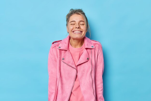 Auténtico concepto de emociones y sentimientos humanos. Feliz joven mantiene los ojos cerrados sonríe con dientes disfruta viendo una película divertida viste una chaqueta rosa aislada sobre fondo azul sonríe a la cámara