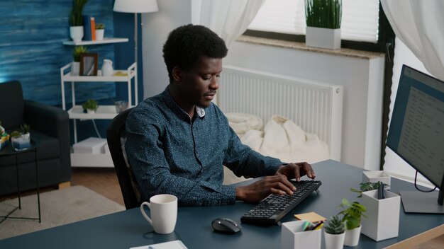 Auténtico hombre afroamericano negro sonriente trabajando desde casa, usuario de computadora de trabajo remoto. Toma de cámara lenta de freelancer mediante comunicación en línea web de internet en piso moderno