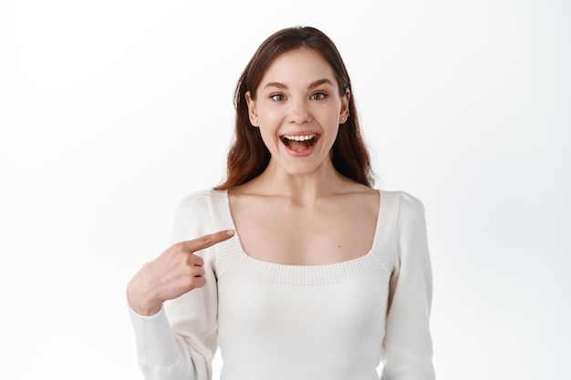 Auto confianza. Joven mujer emocionada señalando con el dedo a sí misma y sonriendo feliz, elígeme gesto, autopromoción, de pie sobre fondo blanco.