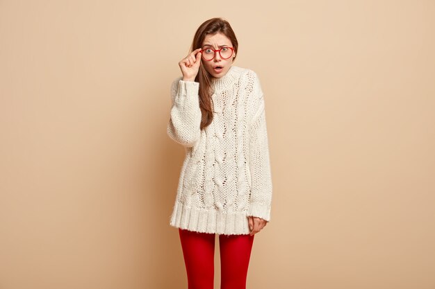 Avergonzada joven modelo mantiene la mano en el borde de las gafas, tiene una expresión facial desconcertada, se sorprende al notar algo increíble e increíble, usa un jersey de invierno, aislado en una pared beige