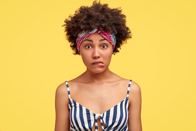 Avergonzada mujer afroamericana frunce los labios, mira con perplejidad, oye algo que no está claro, vestida con un top casual de marinero, aislado sobre una pared amarilla. Personas y expresiones faciales.