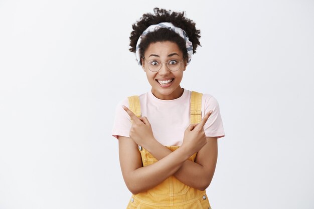 Ayúdame a elegir la dirección. Retrato de mujer inconformista guapa confundida y emocionada con un mono amarillo, diadema y gafas, apuntando a izquierda y derecha con los brazos cruzados y sonriendo con alegría