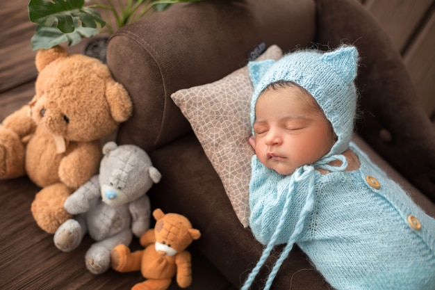 Baby Boy bebé recién nacido durmiendo en pijamas de ganchillo azul en el sofá marrón rodeado de tres osos de juguete