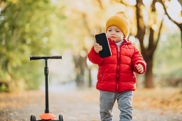 Baby Boy en scooter sosteniendo teléfono móvil en el parque otoñal