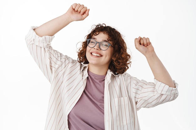 Foto gratuita bailando chica natural divirtiéndose, levantando las manos y de fiesta, sonriendo despreocupada con los ojos cerrados, relajándose en ropa casual, de pie en blanco