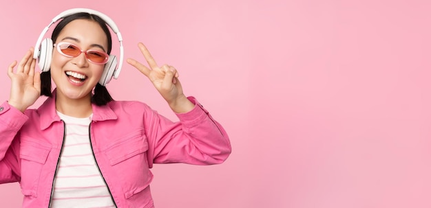 Bailando con estilo chica asiática escuchando música en auriculares posando sobre fondo rosa