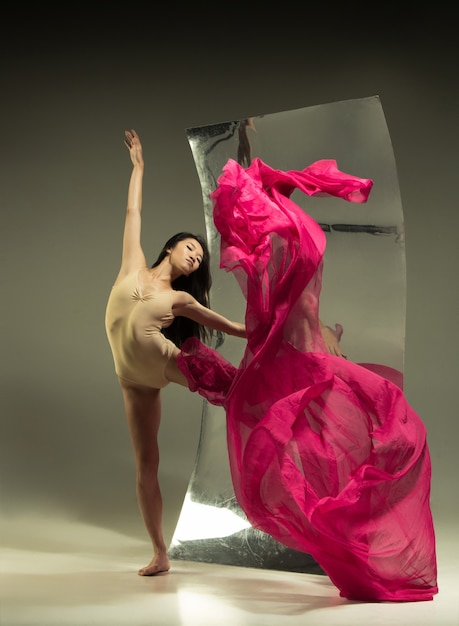 Bailarina de ballet moderno joven y elegante en pared marrón con espejo. Reflejos de ilusión en la superficie. Magia de flexibilidad, movimiento con tela. Concepto de baile de arte creativo, acción, inspirador.