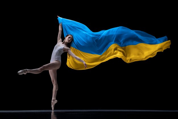 Bailarina de niña bailando con tela pintada en colores azul y amarillo de la bandera de Ucrania en el fondo oscuro del estudio