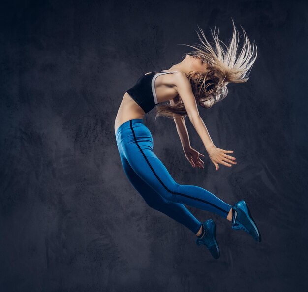 Bailarina rubia joven en ropa deportiva baila y salta en un estudio. Aislado en un fondo oscuro.