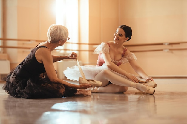 Bailarina sonriente preparándose para la clase con su maestra y poniéndose bombas de ballet mientras se sienta en el suelo