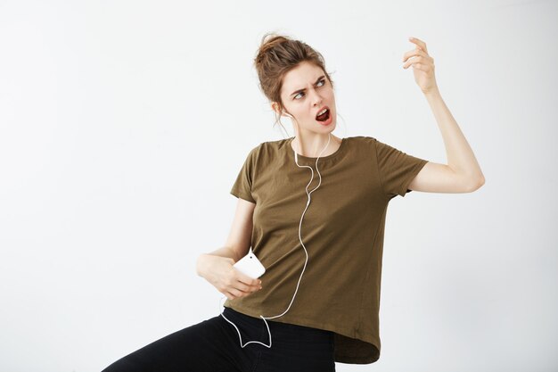 Baile alegre loco de la mujer joven que canta la música que escucha en auriculares sobre el fondo blanco.