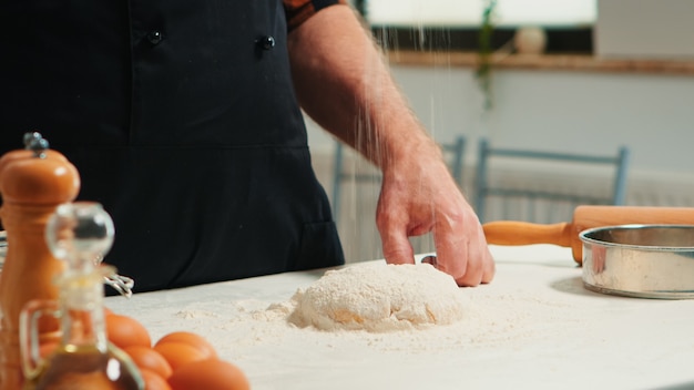 Baker agregando harina en el montón a mano preparando la masa de pan. Cerca del chef anciano jubilado con bonete y rociado uniforme, tamizado y esparcimiento de ingredientes rew para hornear pizzas caseras y cackes.