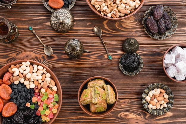Foto gratuita baklava de postre turco de ramadan; lukum; fechas; frutos secos y nueces en cuencos de barro y metálicos contra el escritorio de madera
