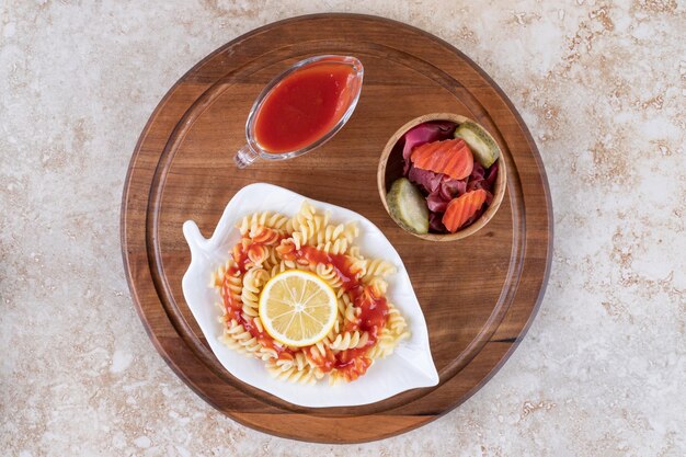 Bandeja de madera debajo de un plato de pasta cocida, ketchup y un cuenco de pepinillos mezclados sobre superficie de mármol.