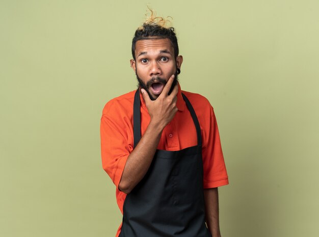 Barbero afroamericano joven sorprendido vistiendo uniforme manteniendo la mano en la barbilla