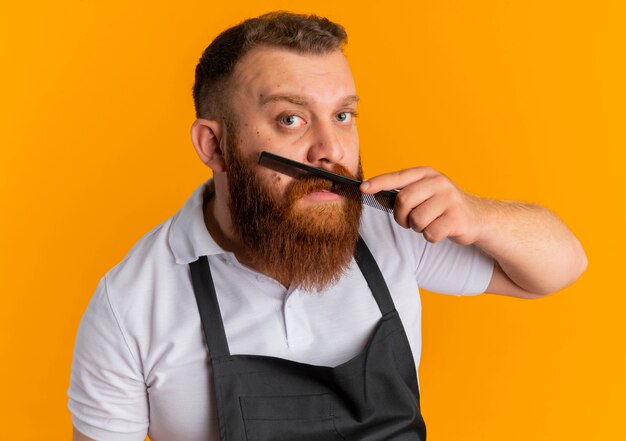 Barbero barbero profesional en delantal peinando su barba de pie sobre la pared naranja