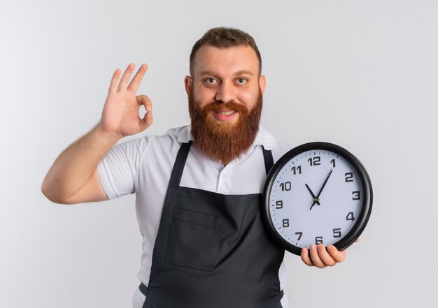 Barbero barbudo profesional hombre en delantal sosteniendo reloj de pared sonriendo mostrando signo ok de pie sobre la pared blanca