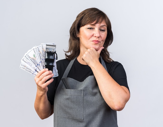 Foto gratuita barbero femenino caucásico adulto confiado en uniforme que sostiene la cortadora de pelo con dinero y mantiene la barbilla aislada sobre fondo blanco con espacio de copia