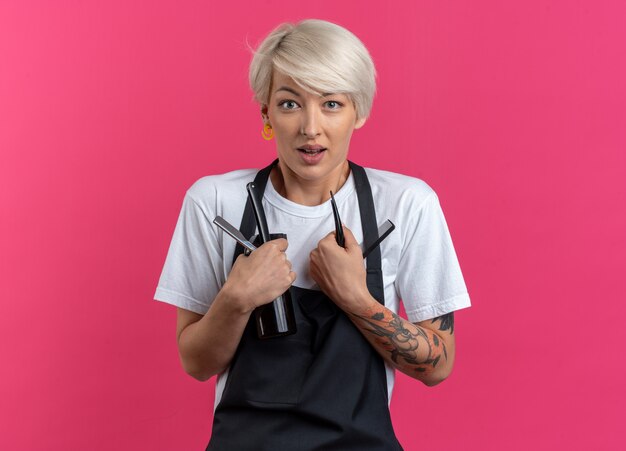 Barbero femenino hermoso joven sorprendido en uniforme que sostiene las herramientas del peluquero aisladas en fondo rosado