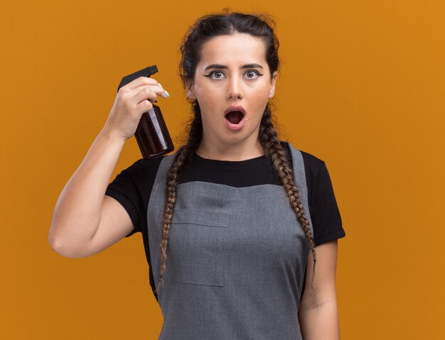 Barbero femenino joven sorprendido en uniforme que sostiene la botella de spray aislada en la pared naranja