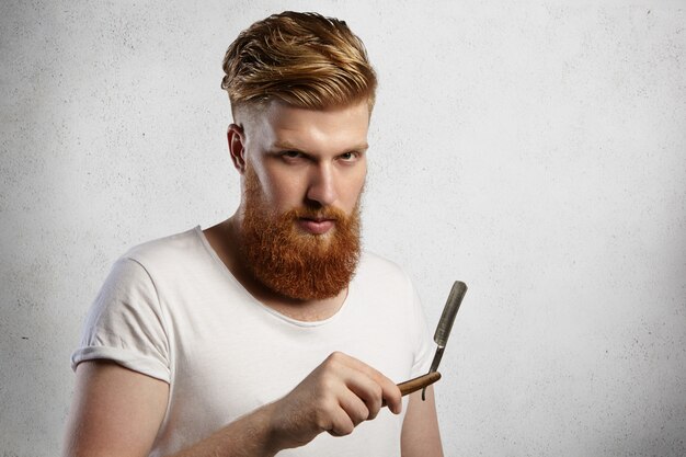 Barbero guapo con barba espesa sosteniendo su accesorio de peluquería, demostrando una afilada hoja de navaja de afeitar.