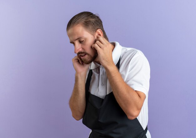 Barbero guapo joven disgustado vistiendo uniforme poniendo los dedos en los oídos con los ojos cerrados aislado sobre fondo púrpura con espacio de copia