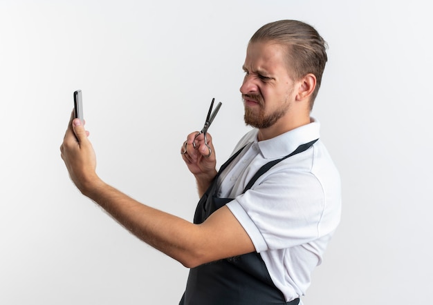 Foto gratuita barbero guapo joven disgustado vistiendo uniforme sosteniendo y mirando el teléfono móvil con unas tijeras en la mano aislado sobre fondo blanco.