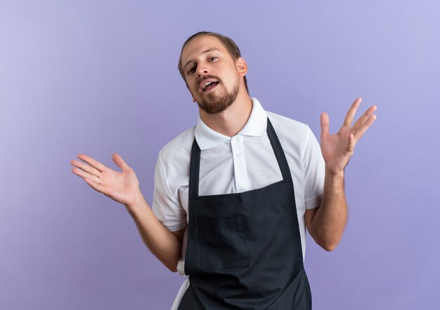 Foto gratuita barbero guapo joven impresionado mostrando las manos vacías con uniforme aislado en púrpura