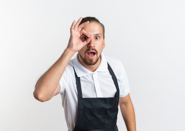 Barbero guapo joven impresionado con uniforme haciendo gesto de mirada aislado en blanco con espacio de copia