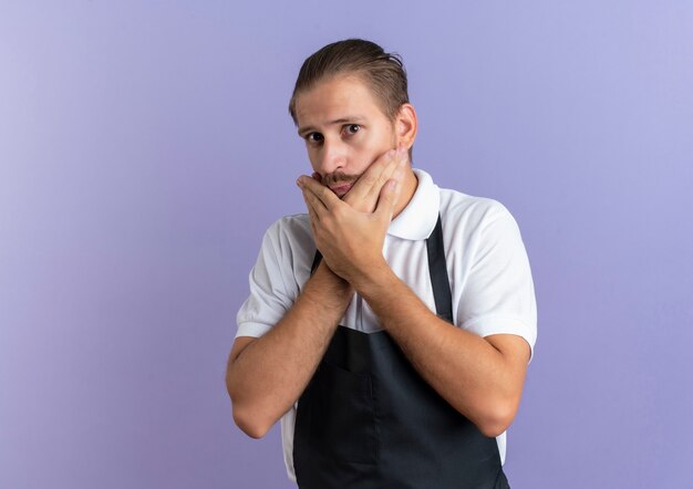 Barbero guapo joven impresionado con uniforme poniendo las manos en el mentón aislado en púrpura con espacio de copia