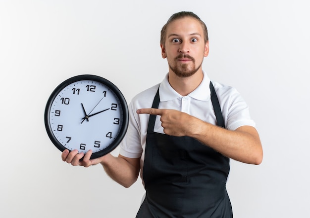 Barbero guapo joven impresionado con uniforme sosteniendo y apuntando al reloj aislado en blanco